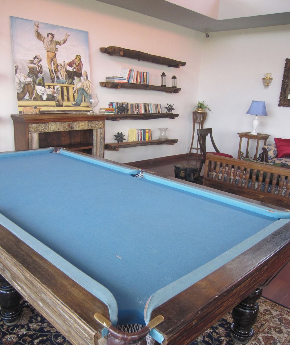 A pool table with a blue felt surface, surrounded by bookshelves, a painting, and a lamp.