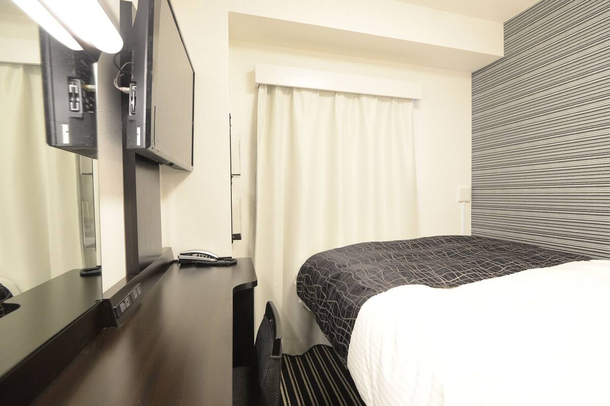 A hotel room with a bed, a desk with a telephone, a mirror, and a window with curtains.
