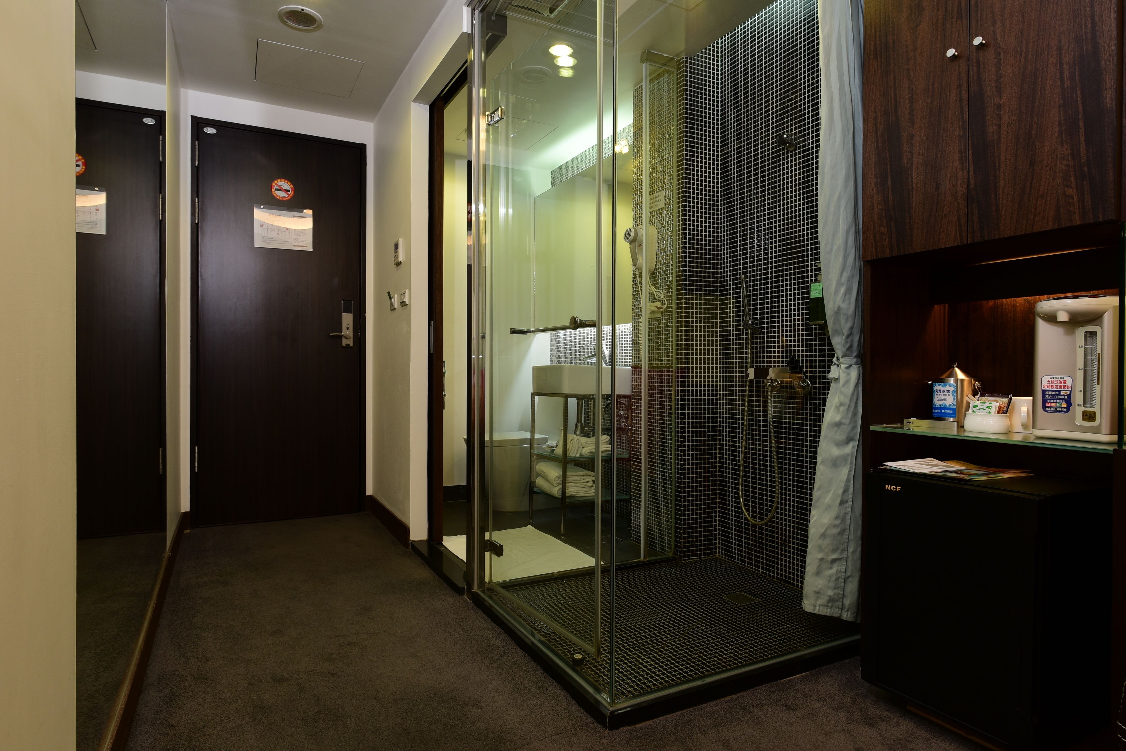 A hotel room with a glass shower enclosure, a small refrigerator, and a wooden cabinet.