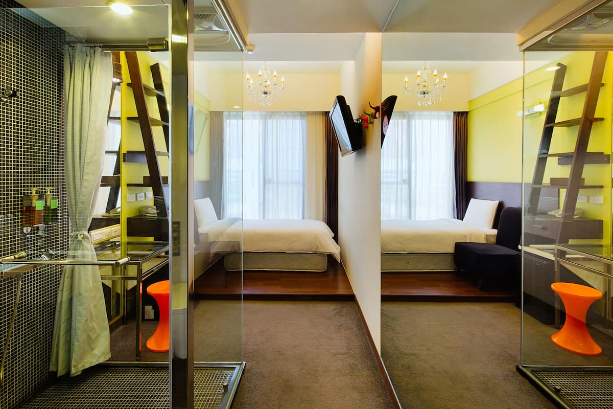 Habitación de hotel con separador de vidrio, dos camas, televisor, candelabro y baño con ducha e inodoro.