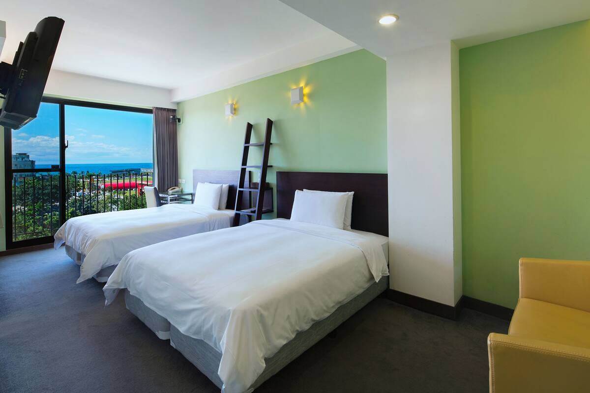 Habitación de hotel con dos camas, un televisor, un balcón con vistas y una silla.