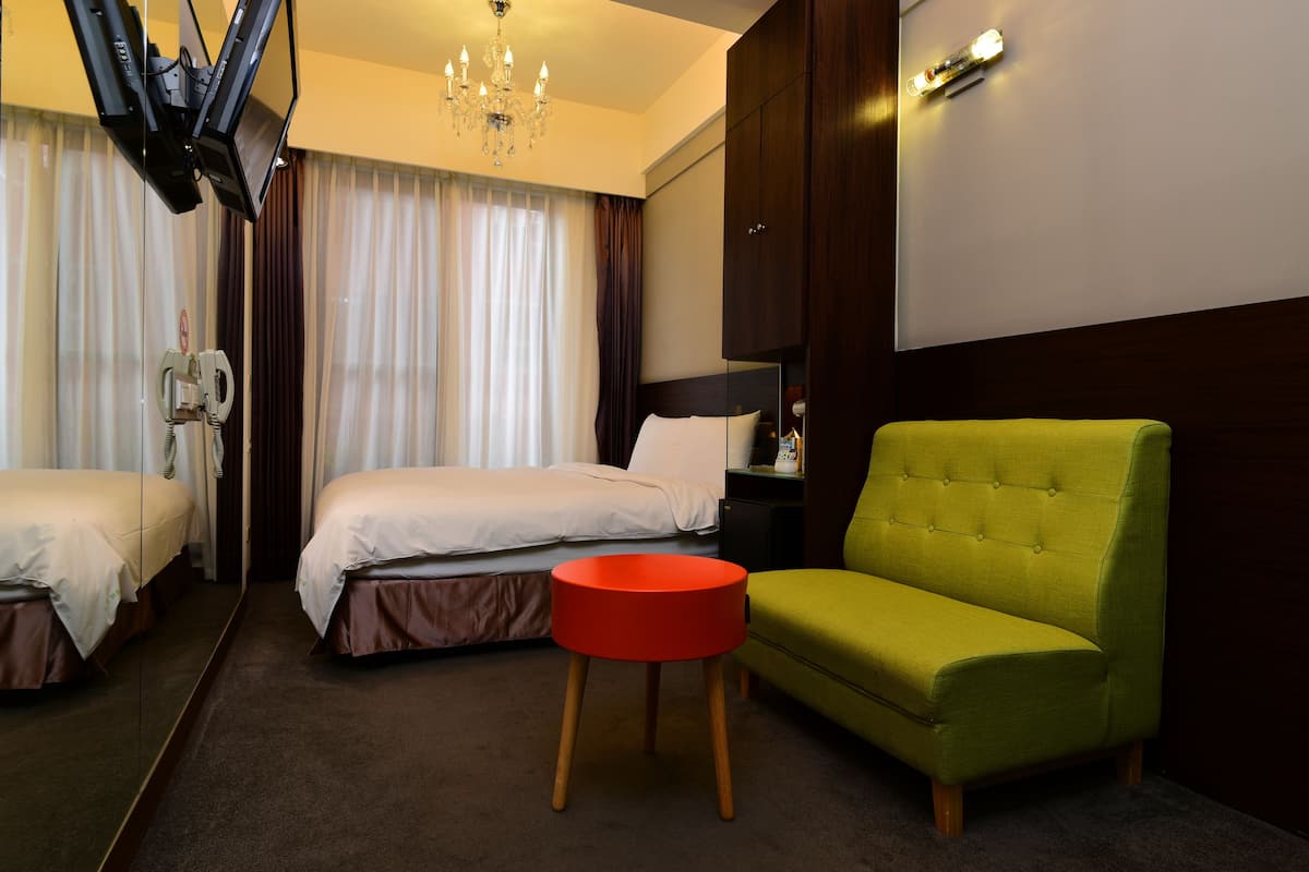 Habitación de hotel con dos camas, un sillón verde, un taburete rojo, una araña y un espejo grande.