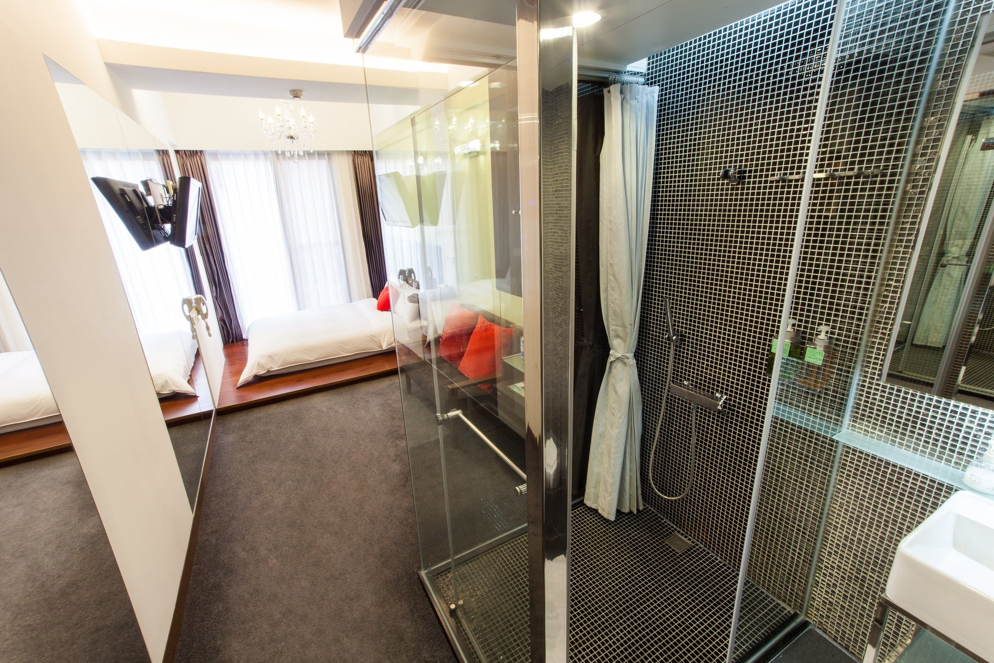A modern hotel room with a glass partition separating the bedroom from the bathroom. The room features a bed, a small seating area, and a shower with a glass enclosure.