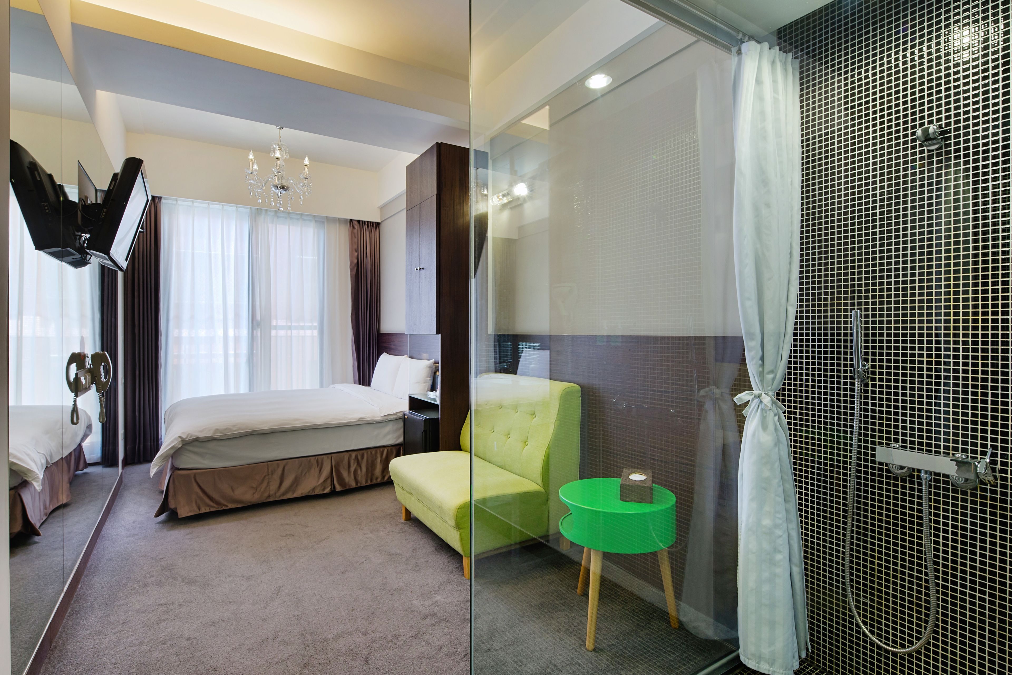 A hotel room with a bed, a green armchair, a small green table, a shower area, and a glass partition.