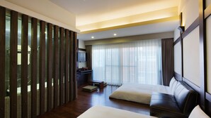 Habitación de hotel con una cama grande, un sofá, un escritorio y vistas de la ciudad a través de amplios ventanales.