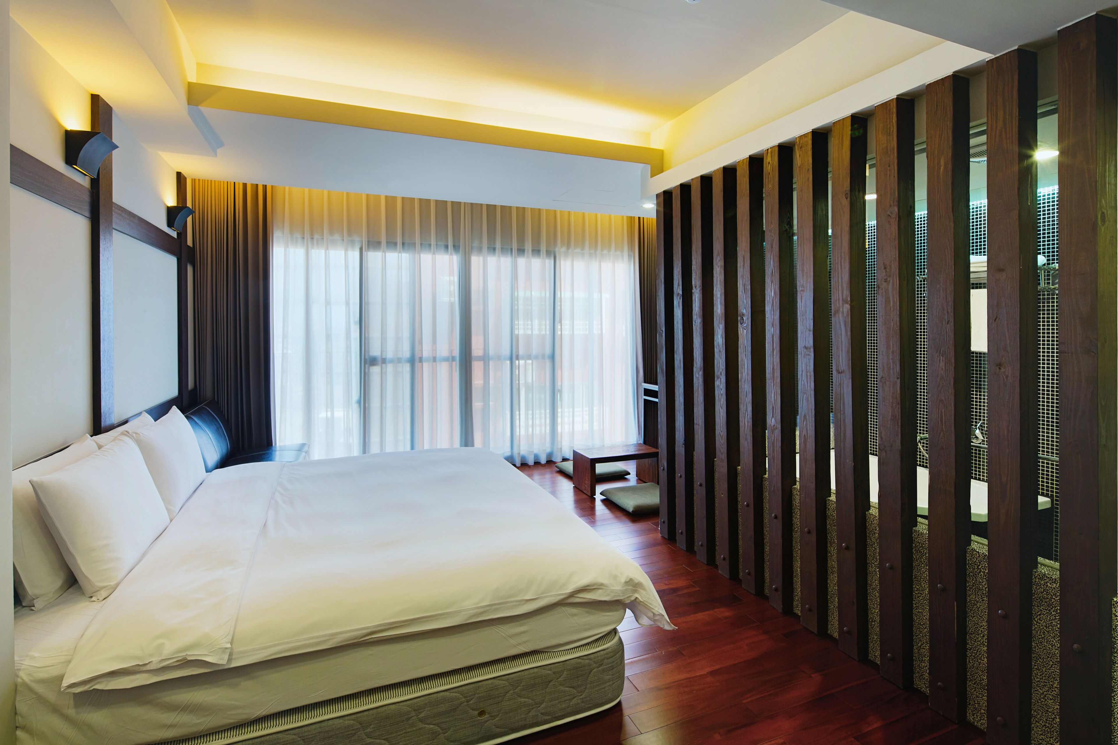 Habitación de hotel moderna con una cama grande, suelo de madera y vistas a través de una ventana con cortinas translúcidas.