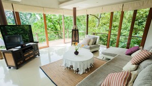 Amplia sala de estar con televisor grande, asientos cómodos y vistas a exuberante vegetación a través de amplias ventanas.