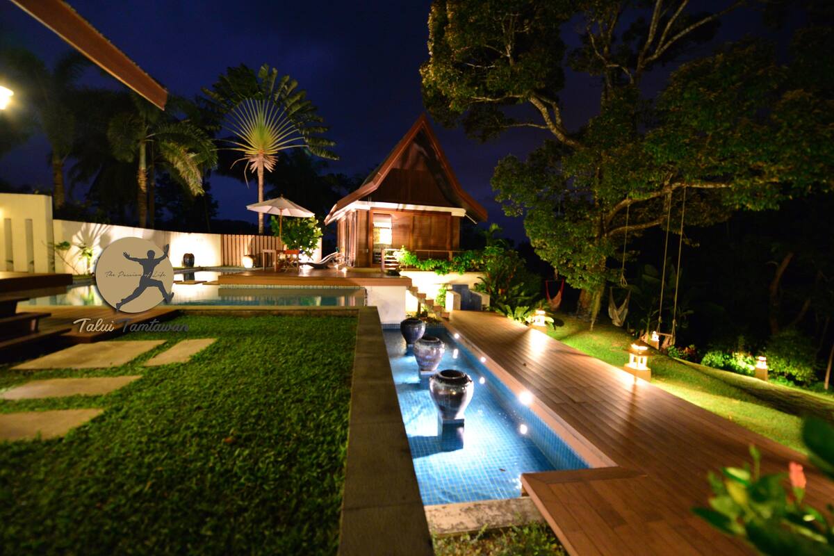Une vue nocturne d’un complexe hôtelier avec une piscine, une terrasse en bois et un petit bâtiment doté d’un toit de chaume.
