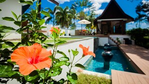Un área de piscina con terraza de madera, un pabellón y plantas tropicales.