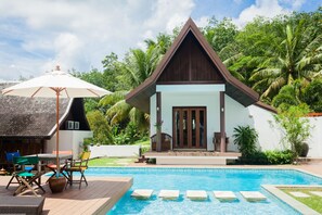 A pool area with a wooden deck, lounge chairs, and a small building with a brown roof.
