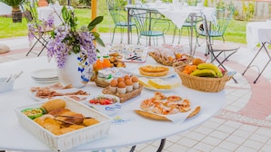 A round table set with various food items, including bread, eggs, cheese, and fruits, in an outdoor setting with green chairs and a white tablecloth. - Hotel Villa Maredona (Ascea)