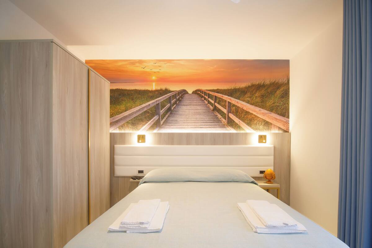 A hotel room with a bed, a nightstand, a lamp, and a painting of a sunset over a boardwalk.