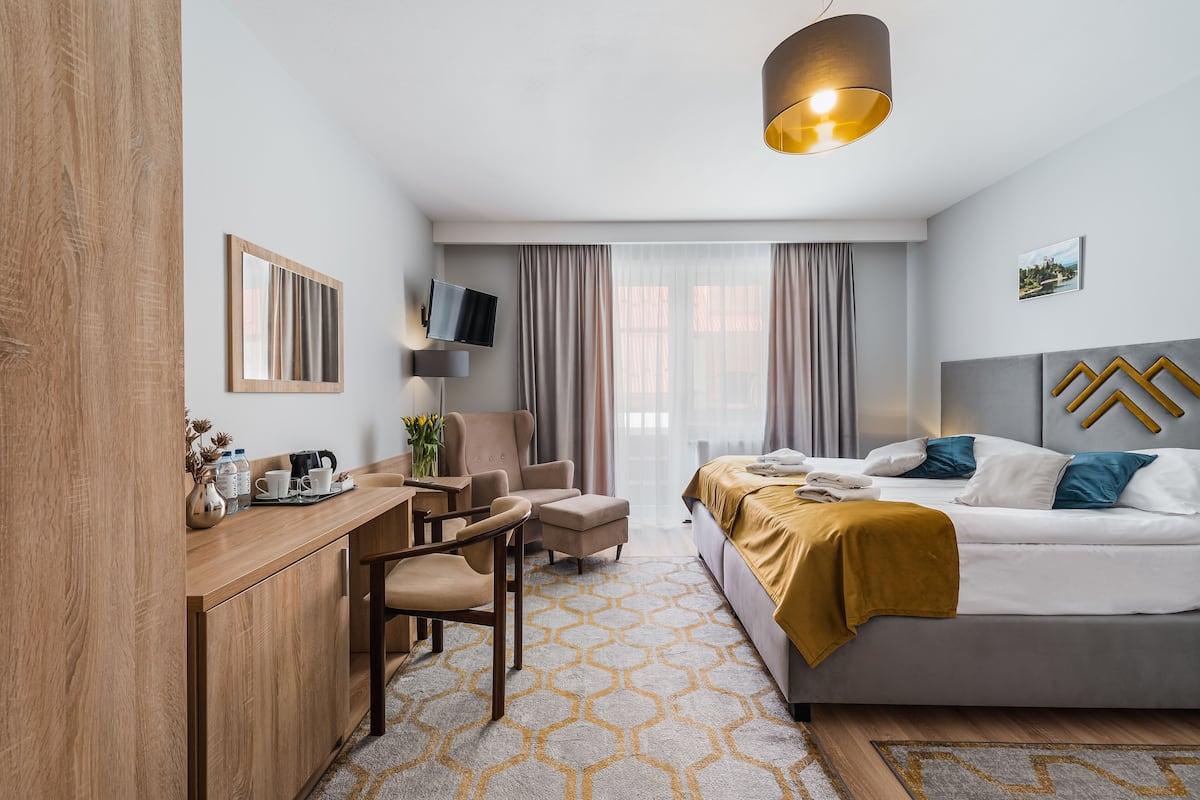 Ein modernes Hotelzimmer mit Schreibtisch aus Holz, einem bequemen Sessel, einer Couch und einem Bett mit goldener Tagesdecke.