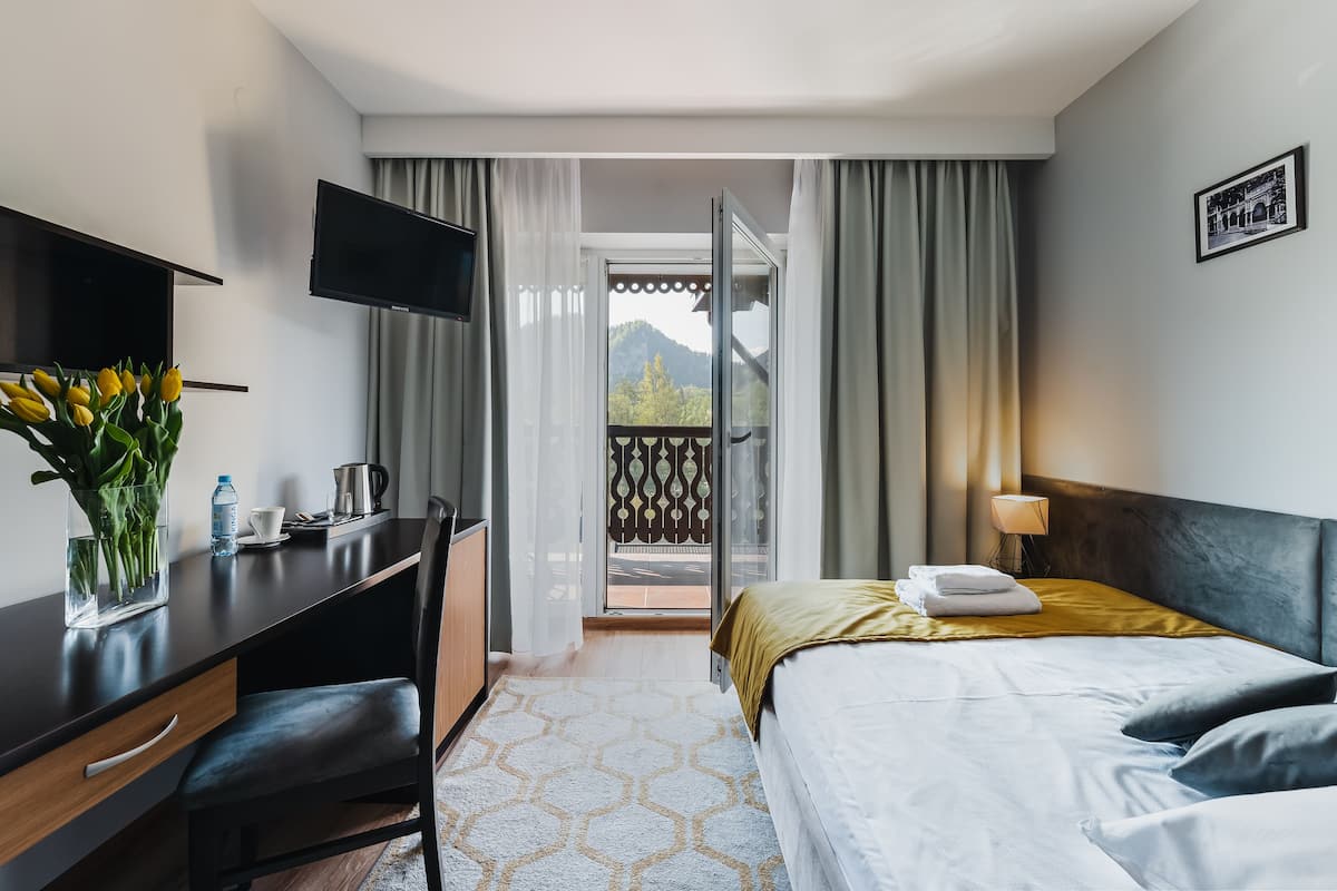 Ein Hotelzimmer mit einem Bett, einem Schreibtisch mit Stuhl, einem Fernseher, einem Fenster mit Vorhängen und einem Balkon mit Blick auf Bäume und Berge.