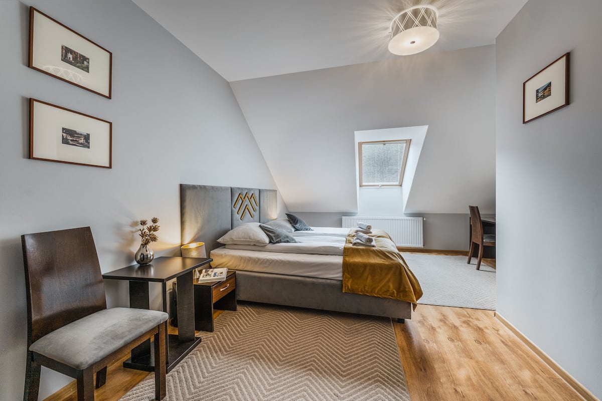 Ein modernes Schlafzimmer mit Bett, Schreibtisch und Stuhl. An der Wand hängen gerahmte Bilder und ein Fenster sorgt für natürliches Licht.