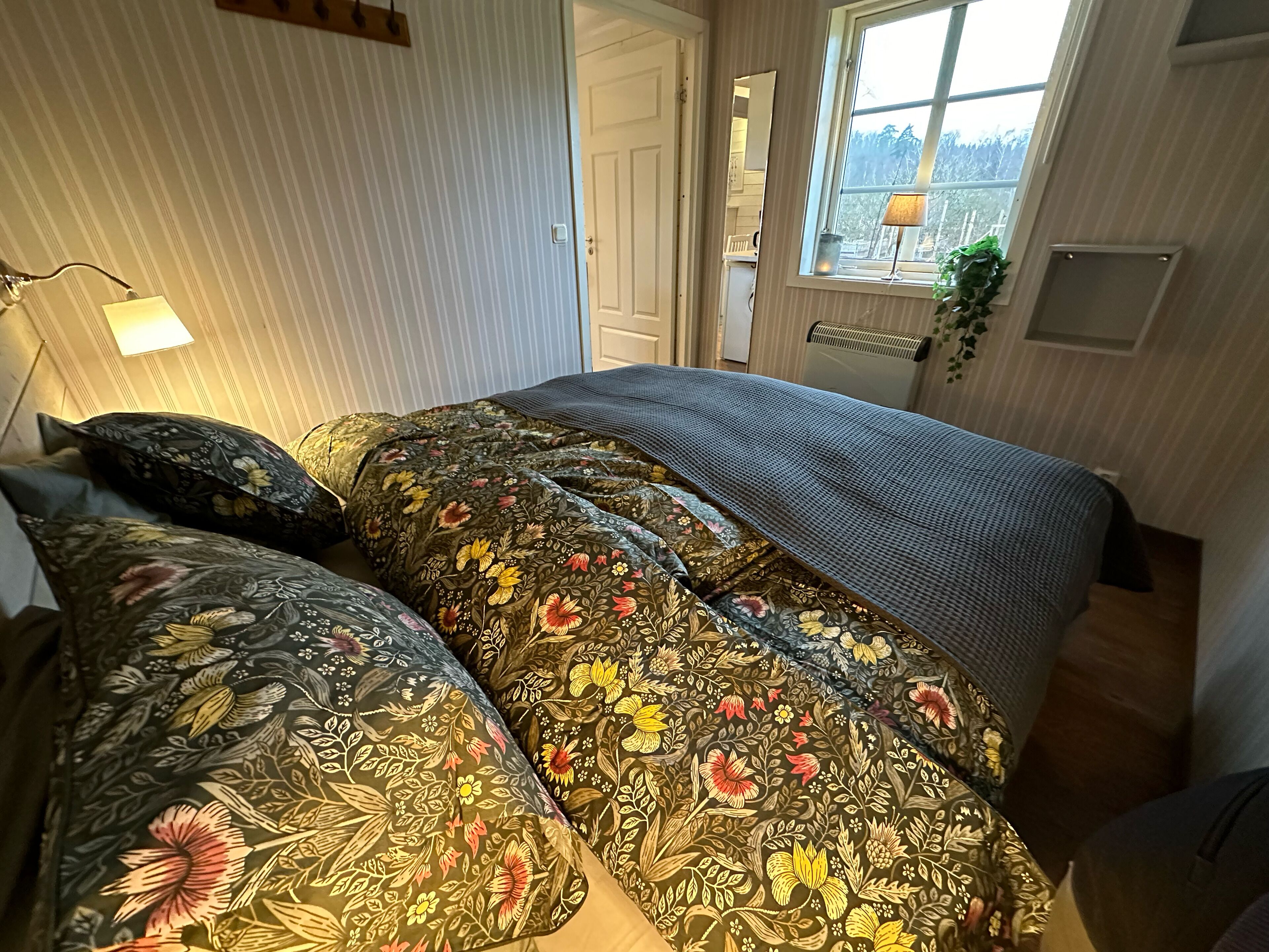 Quarto com cama coberta por roupa de cama estampada com flores, luminária de cabeceira, janela com vista e radiador.