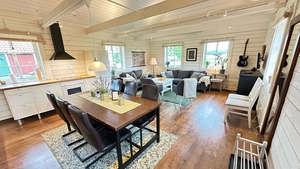 A spacious living area with a wooden dining table, a sofa, and a coffee table. There is a kitchen area with white cabinets and a black stove. - Vindängs Gård (Sloinge)