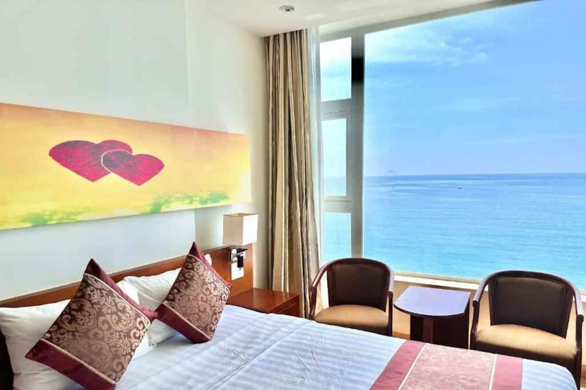 A hotel room with a bed, two chairs, a small table, a painting of hearts, and a large window with a view of the sea.