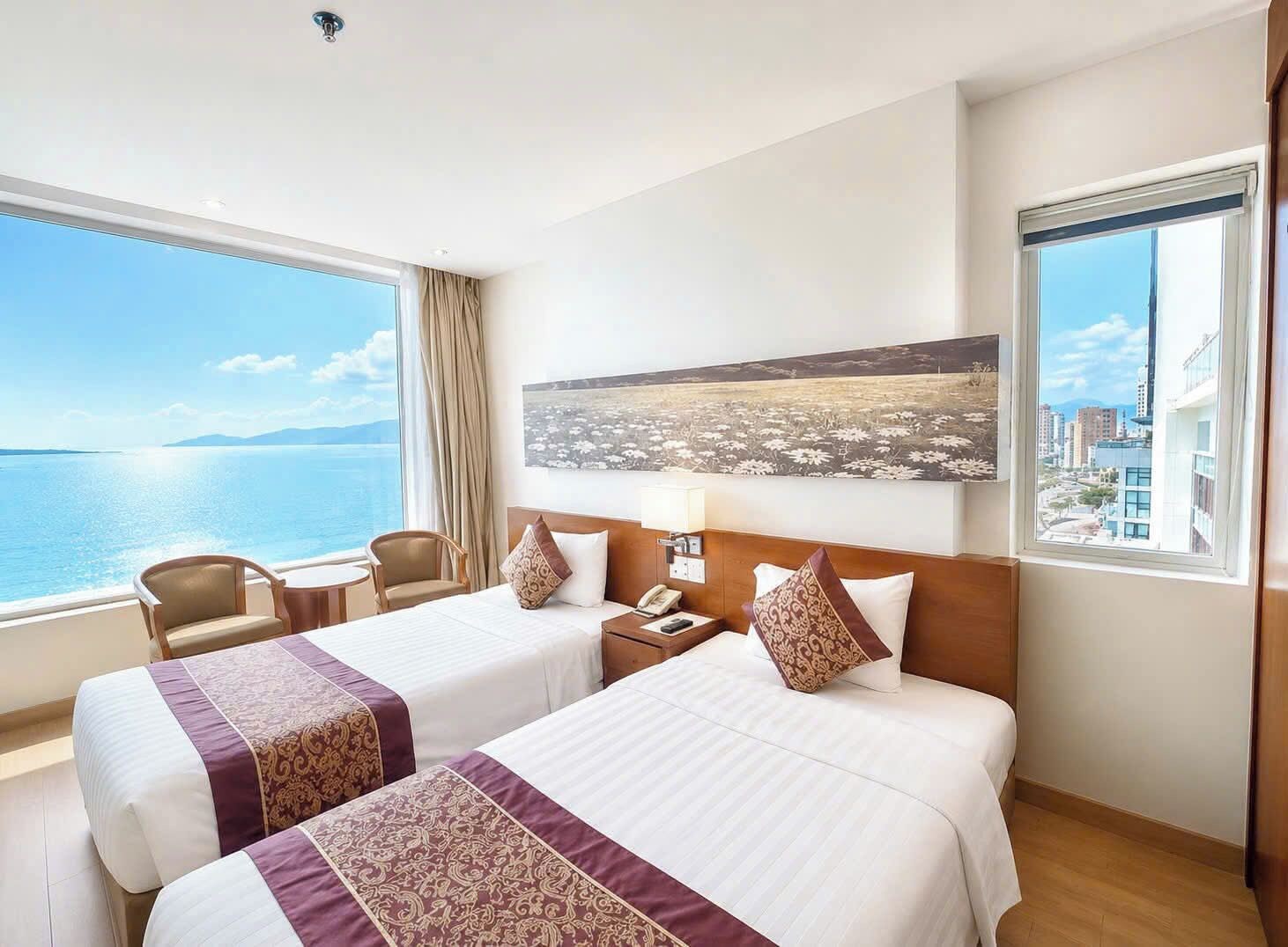 Habitación de hotel con dos camas, un escritorio, una silla, una mesita de noche y un ventanal con vistas al mar y al paisaje urbano.