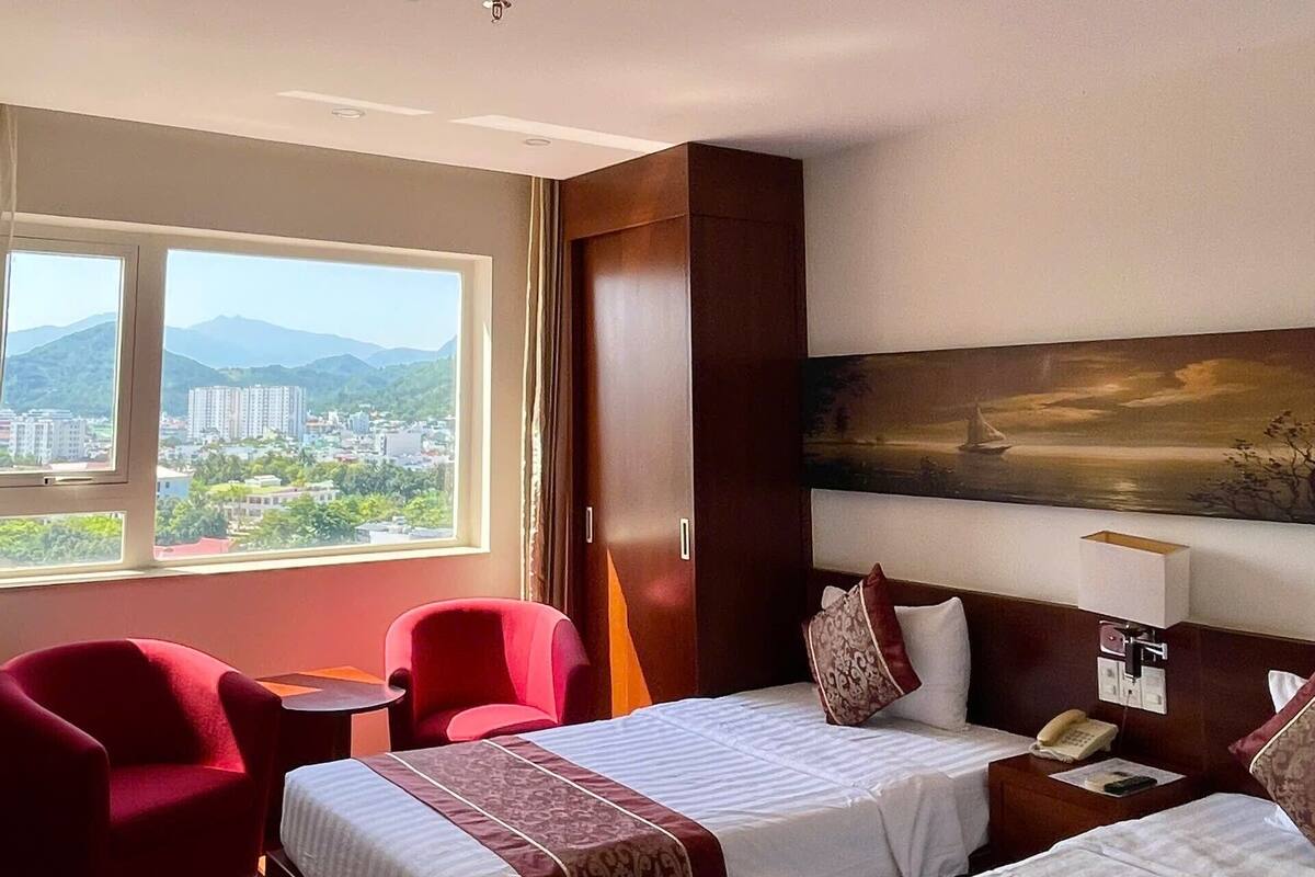 A hotel room with two beds, a red armchair, a small table, a nightstand with a lamp, and a painting on the wall.