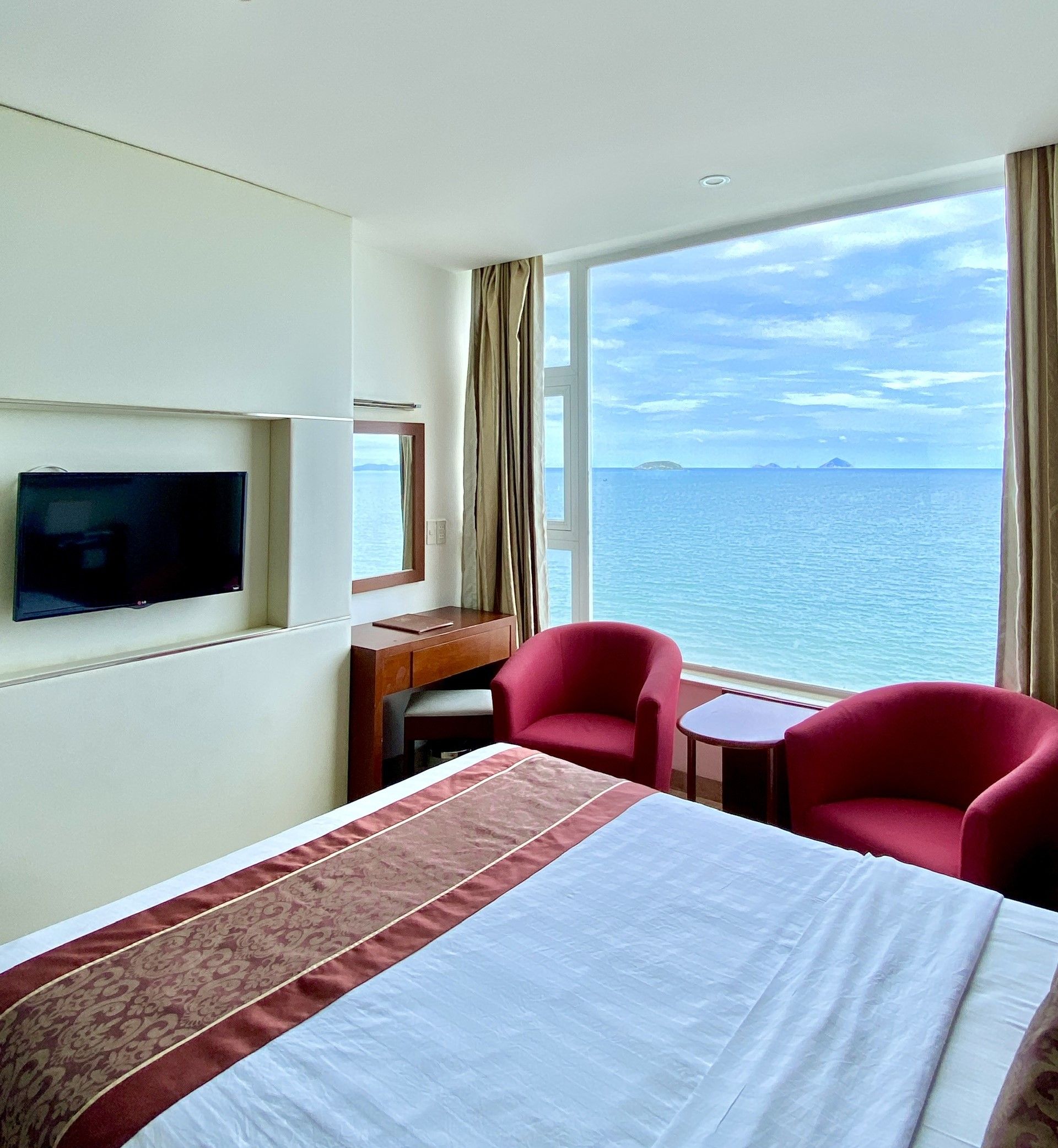 A hotel room with a bed, a desk, a television, and a view of the sea.