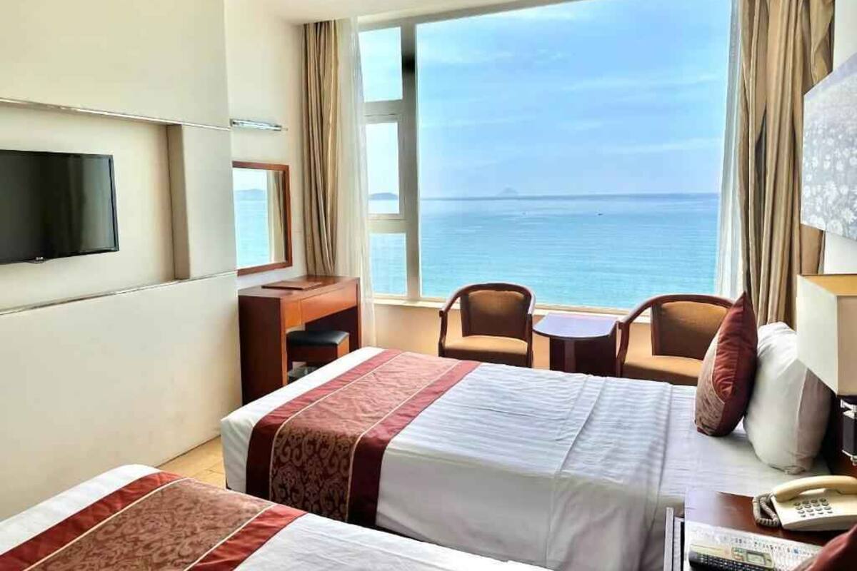 A hotel room with two beds, a TV, a desk, a chair, a table, a phone, and a view of the sea.