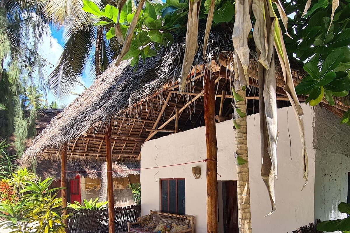 A traditional thatched-roof hut with a red floor, surrounded by lush greenery and palm trees.