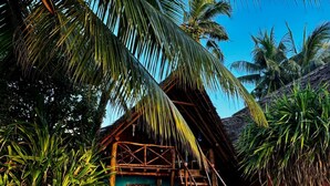 A rustic hut with a thatched roof, surrounded by palm trees and a wooden staircase leading to the entrance. - Evergreen Bungalows (Bwejuu)