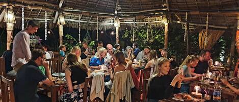 An outdoor dining area with wooden tables and chairs, thatched roof, and hanging lanterns.