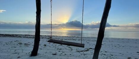 A wooden swing set with a sign reading "Evergreen Bungalows Zanzibar" on a beach with a view of the sea and a sunset.