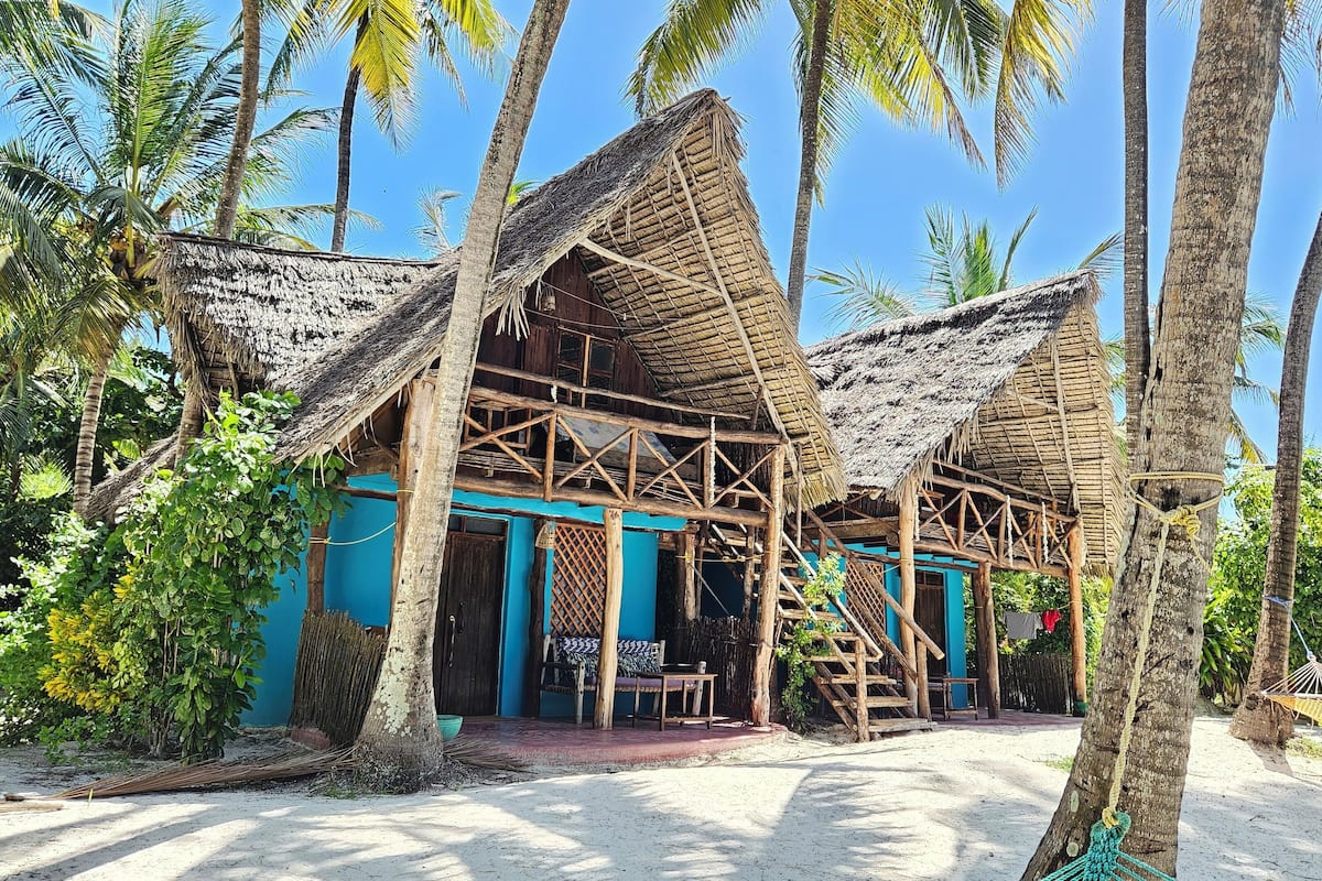 A thatched-roof structure with a blue exterior, surrounded by palm trees and a sandy ground.