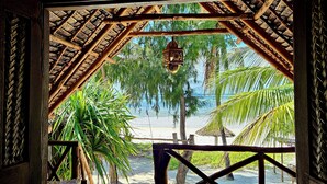 A beach view through a wooden structure with a thatched roof, palm trees, and a hanging lantern. - Evergreen Bungalows (Bwejuu)
