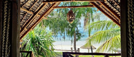 A beach view through a wooden structure with a thatched roof, palm trees, and a hanging lantern.