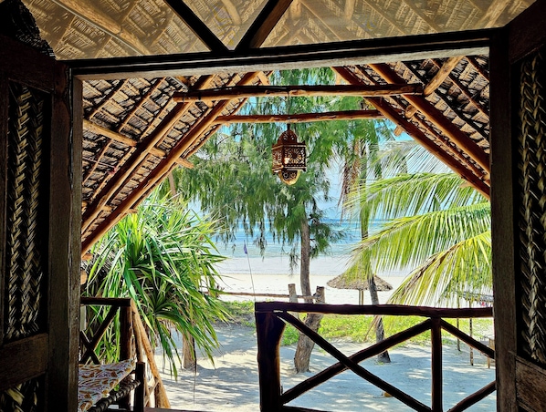 A beach view through a wooden structure with a thatched roof, palm trees, and a hanging lantern. - Evergreen Bungalows (Bwejuu)
