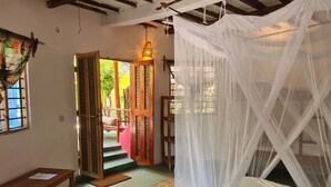 A room with a bed under a white canopy, a wooden bench, a woven mat, and a door leading to an outdoor area with a wicker chair and a lamp. - Evergreen Bungalows (Bwejuu)