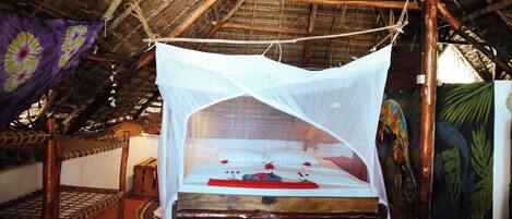 A room with a bed under a mosquito net, wooden furniture, and a thatched roof.