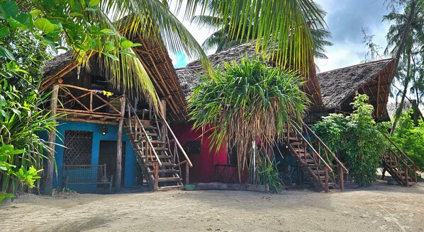 A rustic house with a thatched roof, a blue door, and a wooden staircase leading to the entrance. - Evergreen Bungalows (Bwejuu)