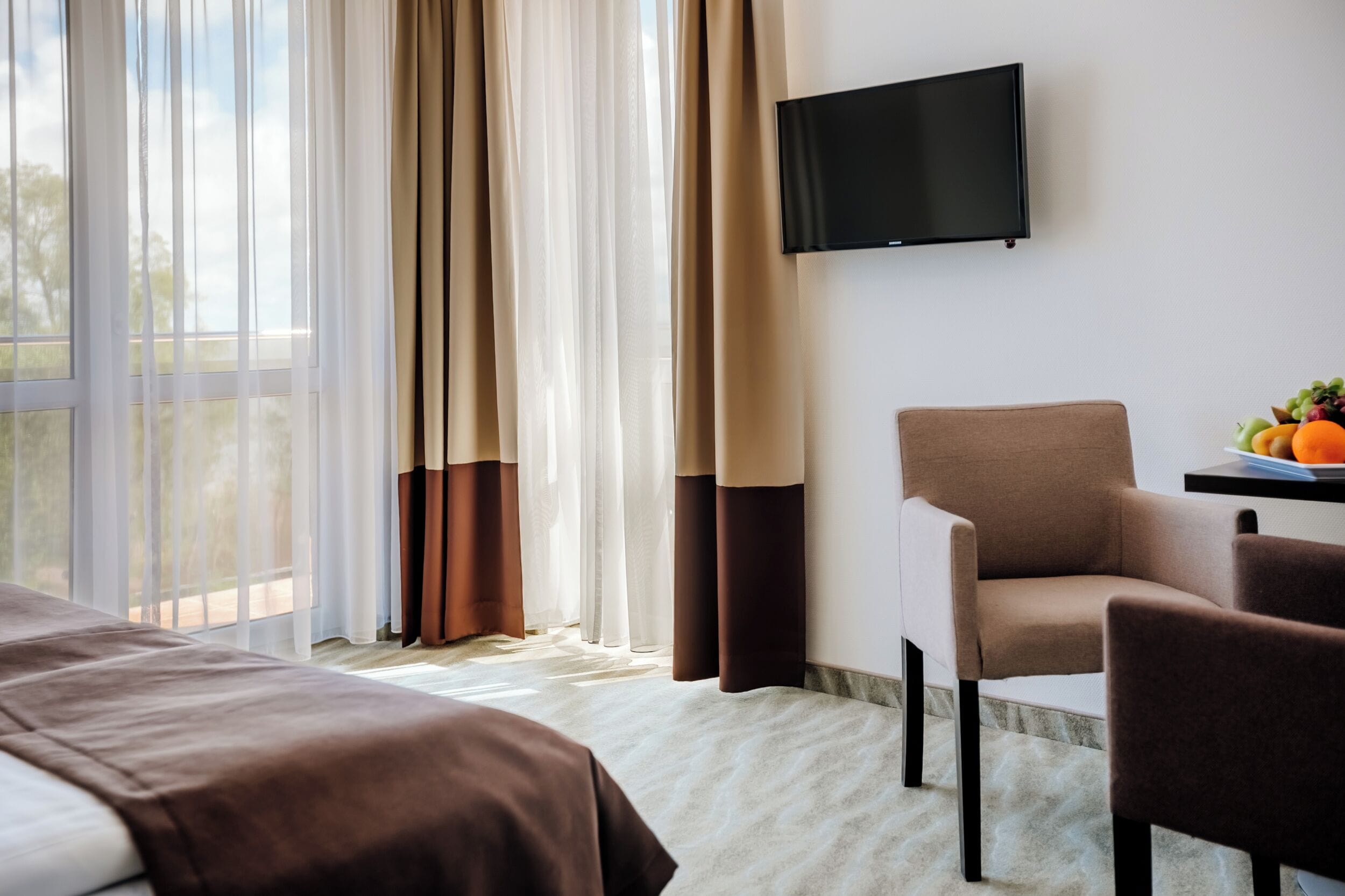 Una camera d'albergo con un letto, una sedia, una televisione a parete e una ciotola di frutta su un tavolo.