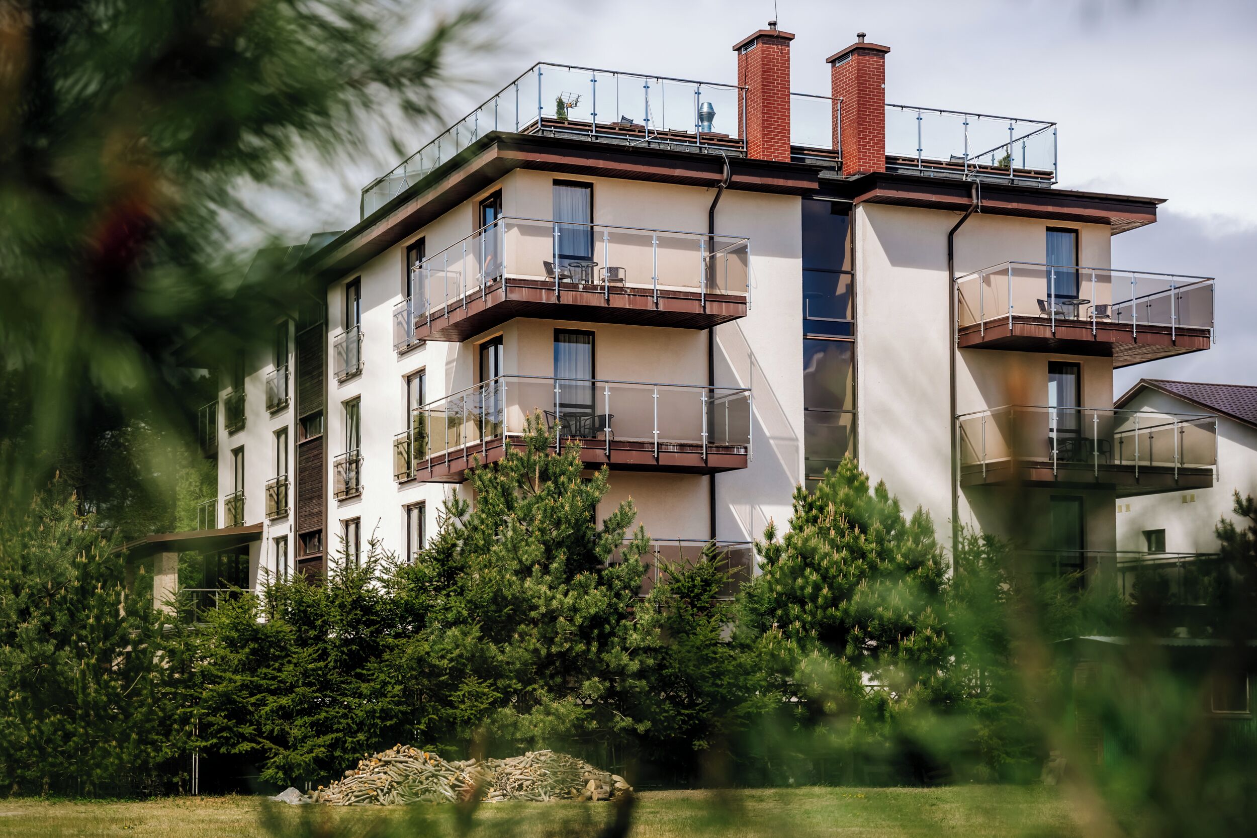 A multi-story residential building with balconies, a brick chimney, and a garden area with trees and a pile of logs.