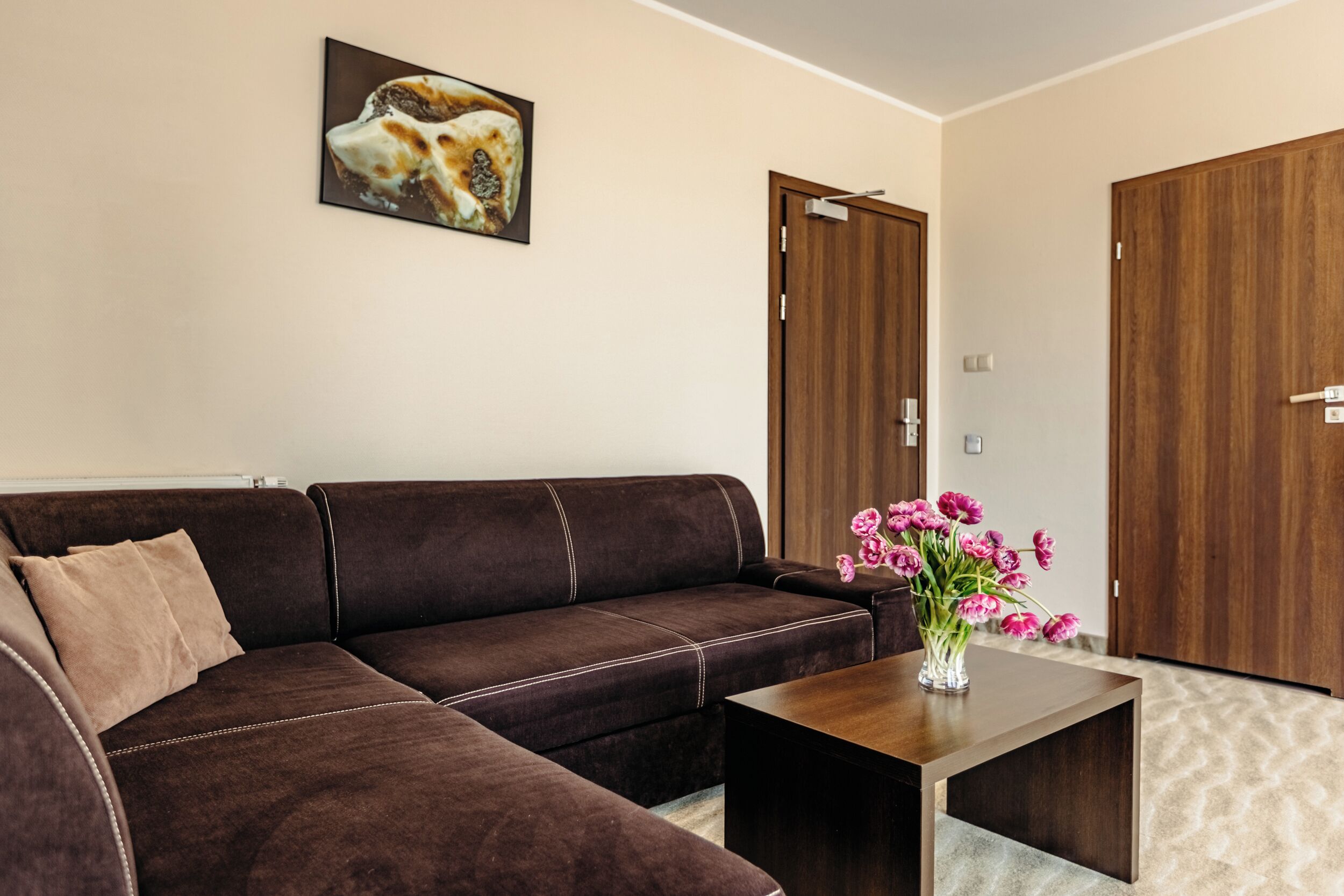 Una sala de estar moderna con un sofá seccional marrón, una mesa de centro de madera con un florero de flores rosas y dos puertas de madera.