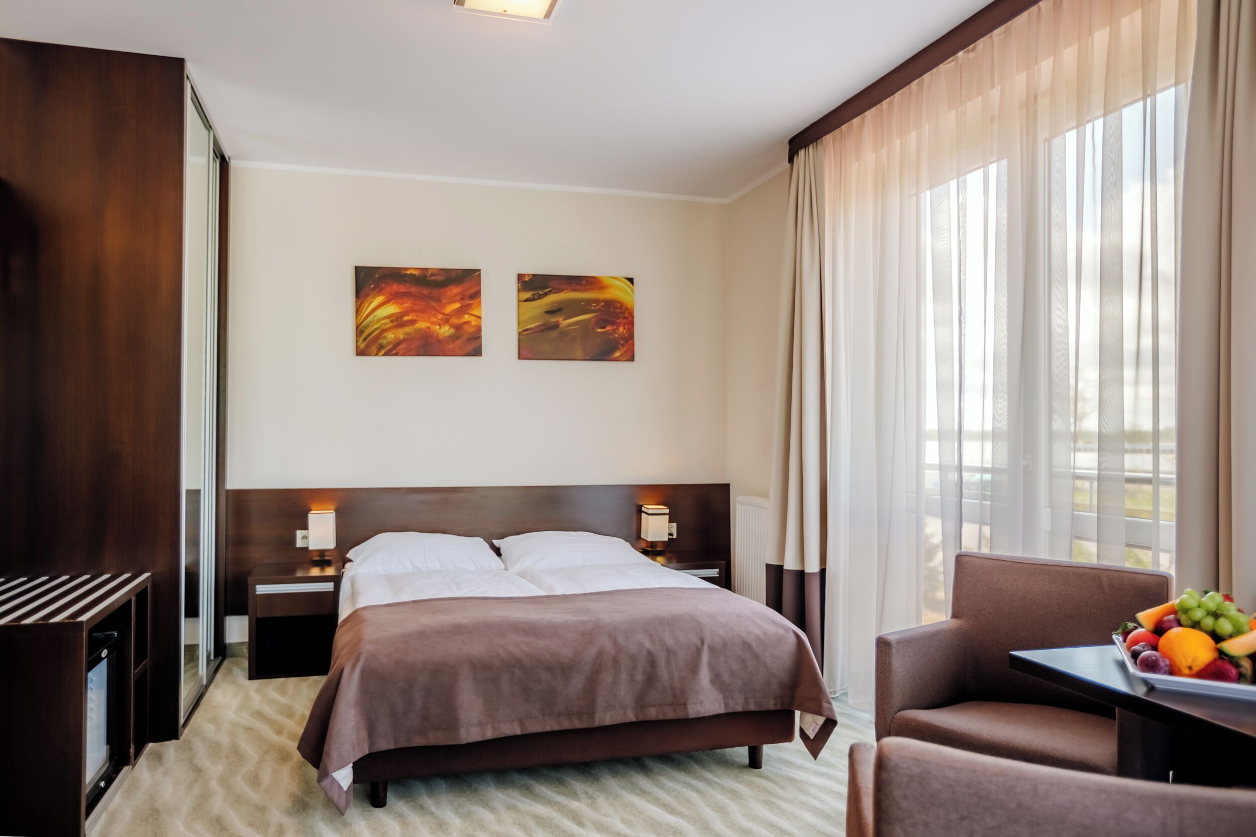 A hotel room with a bed, bedside tables, a desk with a fruit bowl, and a view of the outdoors through sheer curtains.