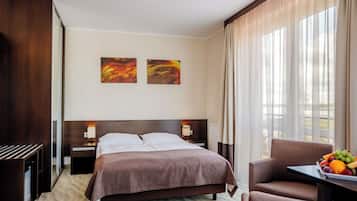 A hotel room with a bed, bedside tables, a desk with a fruit bowl, and a view of the outdoors through sheer curtains.