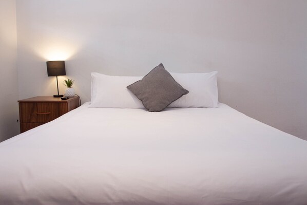 A neatly made bed with white linens and a single gray pillow, a small wooden nightstand with a lamp and a pineapple decoration, and a plain white wall. - Wiley Park Hotel (Wiley Park)