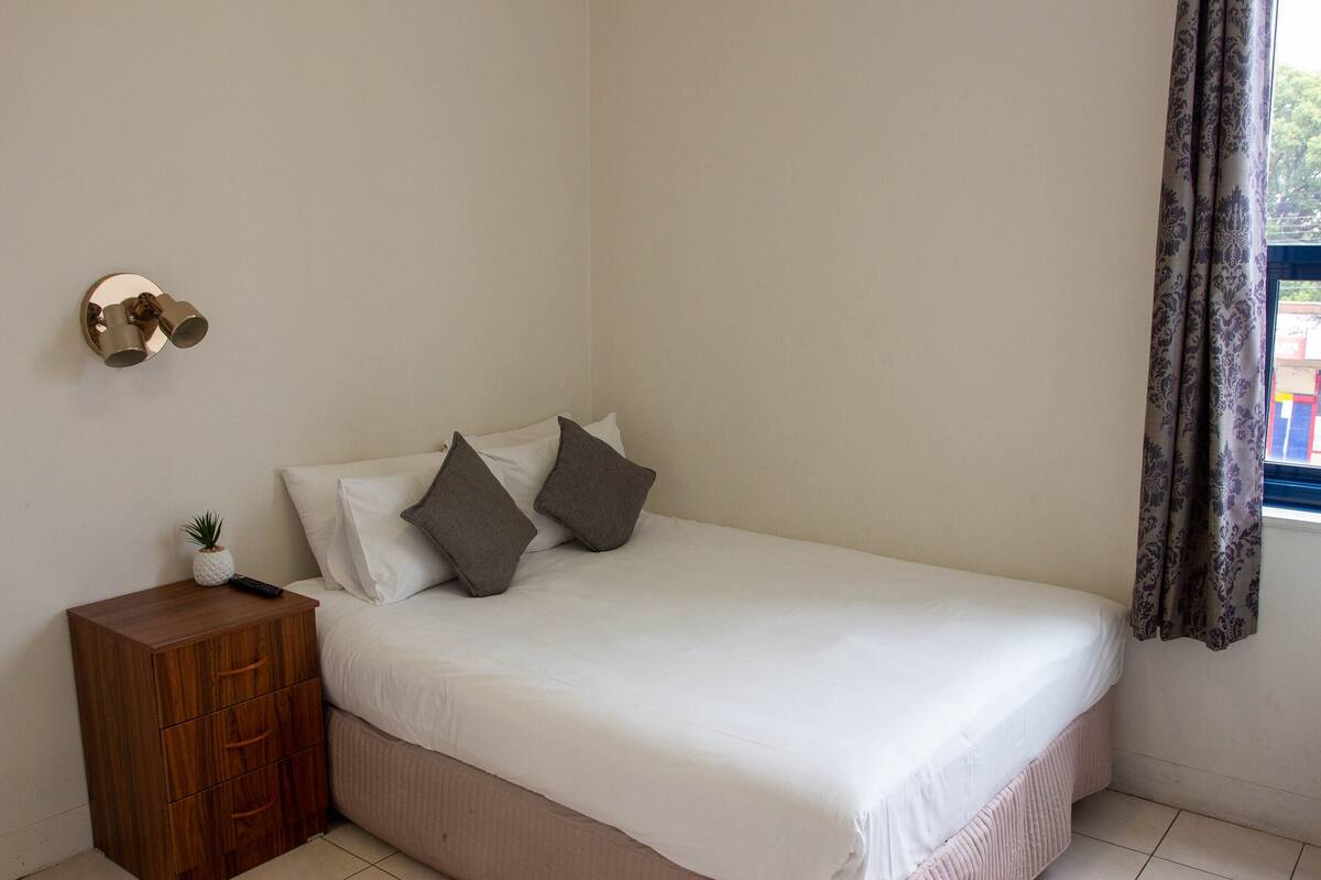 Un dormitorio con cama, mesita de noche, una planta pequeña y una lámpara de pared.