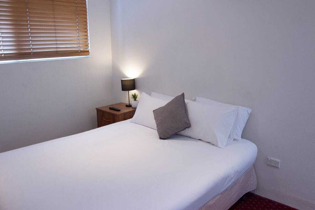Una cama bien hecha con ropa de cama blanca y un cojín gris, una lámpara de noche y una pequeña planta en maceta sobre una mesita de noche de madera.