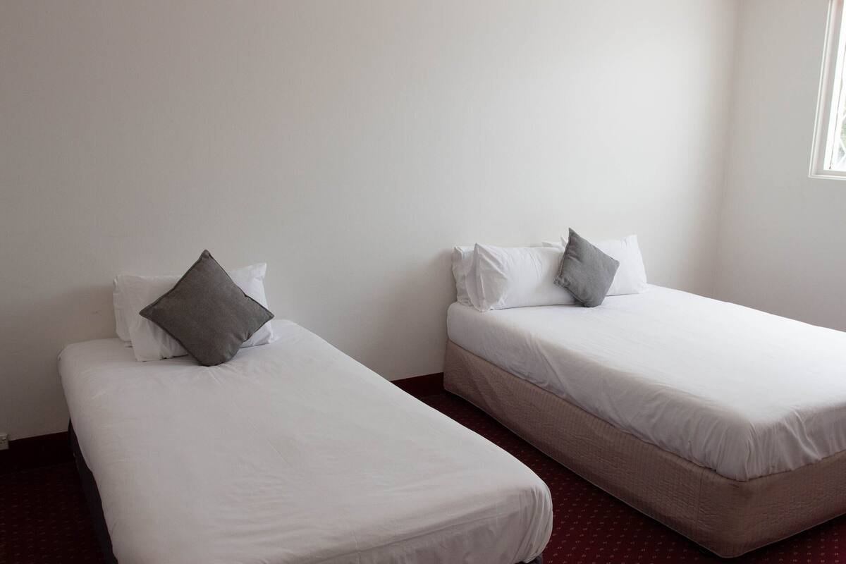Dos camas individuales con sábanas blancas y almohadas grises en una habitación con pared blanca lisa y una ventana a la derecha.