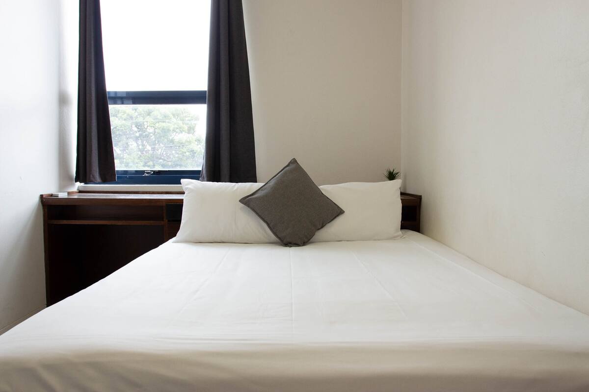 Una cama bien hecha con sábanas blancas y una única almohada gris, ubicada junto a una ventana con cortinas oscuras.