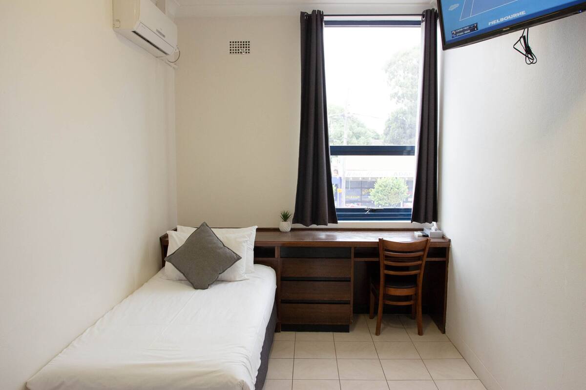 Una habitación pequeña y limpia con una cama, un escritorio y una silla. Hay una ventana con cortinas, un televisor montado en la pared y un aire acondicionado.