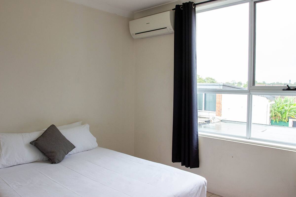 Habitación de hotel con cama, ventana con cortina y unidad de aire acondicionado en la pared.