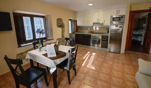 A dining area with a table set for two, a bottle of champagne, and a vase with purple flowers. The kitchen in the background features stainless steel appliances and white cabinets. - Casona de Llanorrozo (Cudillero)